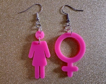 May include: A pair of pink earrings. One earring is shaped like a person and the other is shaped like the female gender symbol.