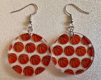 May include: A pair of earrings with a basketball pattern. The earrings are made of clear acrylic and have silver hooks. Each earring features a circle with a repeating pattern of orange and black basketballs.