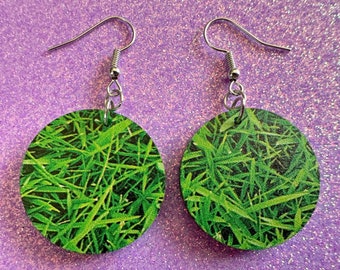 May include: A pair of green grass-themed earrings with silver hooks. The earrings are round and feature a close-up image of green grass.