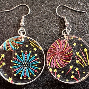 Puede incluir: Un par de pendientes de plata con colgantes redondos transparentes que presentan un diseño de fuegos artificiales. Los fuegos artificiales son en tonos de rojo, amarillo, azul y rosa sobre un fondo negro.