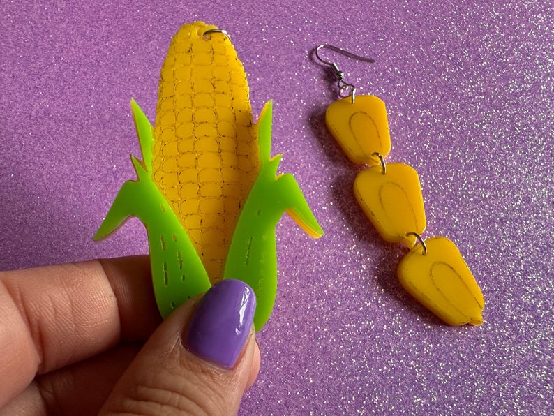 May include: A pair of earrings featuring a corn on the cob design. One earring is a single corn cob with green husks, while the other earring is a chain of three corn kernels.