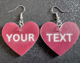 May include: A pair of heart-shaped earrings with silver hooks. The hearts are a deep red color and have white text that reads "YOUR TEXT" on them.