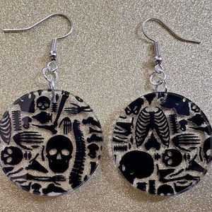 May include: A pair of silver dangle earrings with clear round discs. The discs are decorated with a black and white pattern of skulls and bones.
