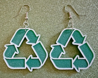 May include: A pair of green and white recycle symbol earrings. The earrings are made of a textured material and have silver hooks.