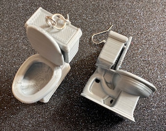 May include: A pair of gray 3D printed toilet earrings. The earrings are shaped like toilets with the lids open. The toilets are attached to silver earring hooks.