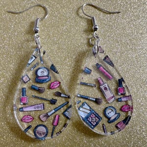 May include: Clear, teardrop-shaped earrings with a silver hook. Each earring is decorated with a variety of makeup items, including lipsticks, brushes, eyeshadow, and nail varnish. The makeup items are in pink, blue, and black.