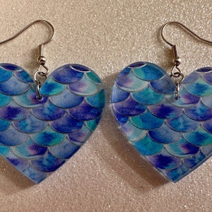 Puede incluir: Un par de pendientes en forma de corazón con un diseño de escamas de sirena en tonos azules, morados y blancos. Los pendientes tienen ganchos plateados.