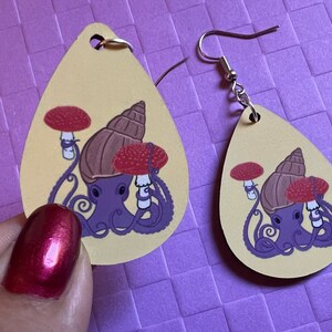May include: A pair of teardrop-shaped earrings with a yellow background. Each earring features a purple octopus with a brown snail shell on its head. The octopus is holding two red and white mushrooms. The earrings have silver hooks.