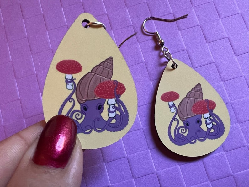 May include: A pair of teardrop-shaped earrings with a yellow background. Each earring features a purple octopus with a brown snail shell on its head. The octopus is holding two red and white mushrooms. The earrings have silver hooks.
