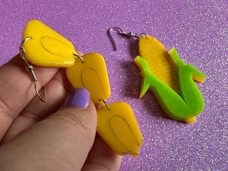 May include: A pair of yellow corn cob earrings with silver hooks. The earrings are made of acrylic and are shaped like corn cobs. The earrings are a fun and quirky way to add a touch of personality to any outfit.
