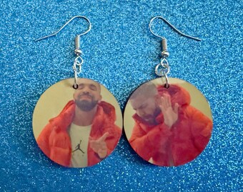 May include: A pair of round earrings featuring a meme of a man in a red jacket. The earrings have silver-colored hooks. The image on the earrings is a popular internet meme.