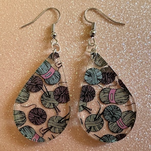 May include: A pair of teardrop-shaped earrings with a clear background and a pattern of colorful yarn balls and knitting needles.