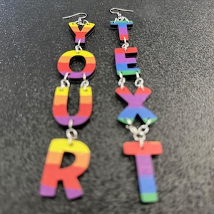 May include: Rainbow-coloured dangle earrings spelling out the words "YOUR NEXT". Each letter is a different colour, with a silver hook at the top. The earrings are suspended from silver chains.