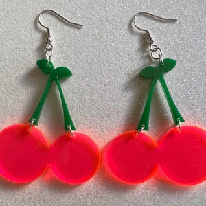 以下が含まれることがあります： 緑色の茎と銀色のフックが付いた、ネオンピンクのチェリーの形をしたイヤリングのペアです。
