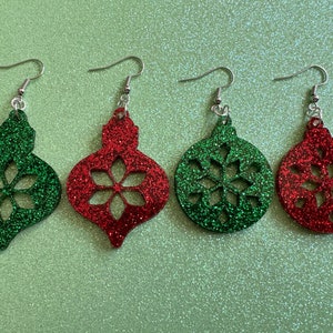 May include: Four pairs of Christmas ornament earrings. Two pairs are red with glitter and a snowflake cutout. Two pairs are green with glitter and a snowflake cutout.
