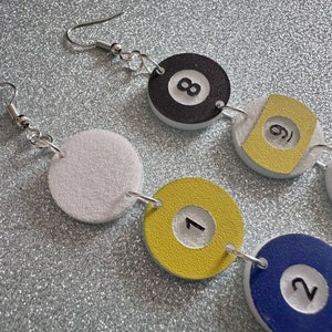 May include: A pair of earrings with a white, yellow, blue, and black color scheme. The earrings feature a series of round charms with numbers 1, 2, 6, 8, and 10 printed on them. The charms are connected by silver jump rings.