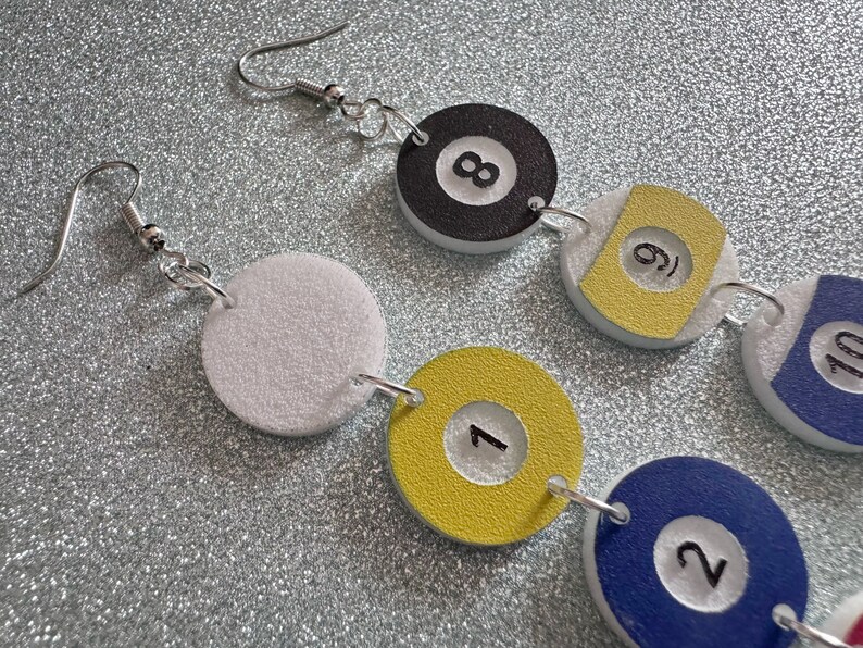May include: A pair of earrings with a white, yellow, blue, and black color scheme. The earrings feature a series of round charms with numbers 1, 2, 6, 8, and 10 printed on them. The charms are connected by silver jump rings.