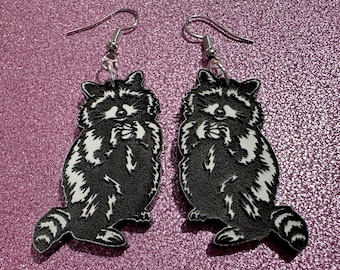 May include: A pair of black and white raccoon earrings with silver hooks. The raccoons are depicted in a sitting position with their paws raised to their faces.