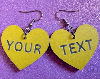 May include: A pair of yellow heart-shaped earrings with silver hooks. The left earring says "YOUR" and the right earring says "TEXT".