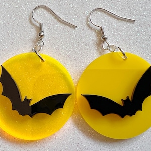 May include: A pair of yellow circle earrings with a black bat silhouette on each. The earrings have silver hooks.