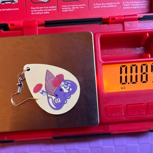 May include: A small, white, ceramic earring with a purple octopus, a red mushroom, and a brown shell design. The earring is on a digital scale that reads 0.085 oz.