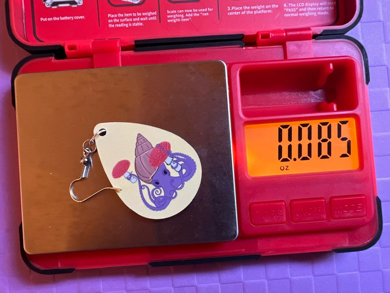 May include: A small, white, ceramic earring with a purple octopus, a red mushroom, and a brown shell design. The earring is on a digital scale that reads 0.085 oz.