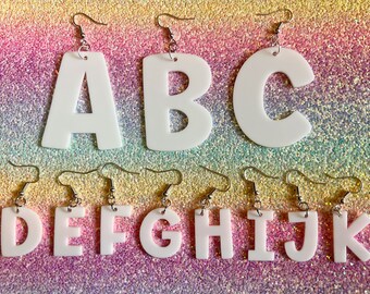 May include: White acrylic letter earrings, spelling out the letters A through K. Each letter is suspended from a silver-colored fish hook earring. The background is a glittery, iridescent mix of pink, purple, and gold.