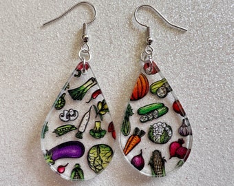 May include: Clear, teardrop-shaped earrings with a variety of colorful vegetable illustrations. The earrings feature a silver hook and are adorned with images of carrots, corn, eggplant, and more.