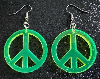 May include: A pair of green peace sign earrings with silver hooks. The peace signs are made of acrylic and have a slightly glittery finish.