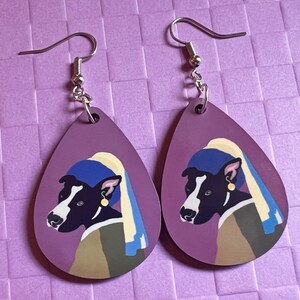 Könnte beinhalten: Ein Paar Hängeohrringe mit einem lila Hintergrund und einem schwarz-weißen Hund, der ein blaues und gelbes Stirnband trägt. Der Hund trägt einen goldenen Ohrring.