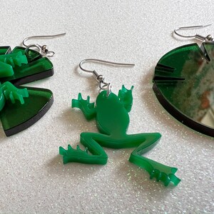 May include: A pair of green acrylic earrings featuring a frog and a lily pad design. The frog is sitting on the lily pad.