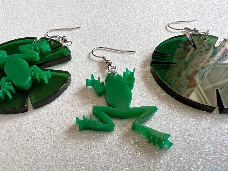 May include: A pair of green acrylic earrings featuring a frog and a lily pad design. The frog is sitting on the lily pad.