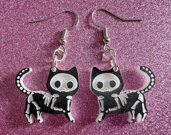 May include: A pair of black and white skeleton cat earrings with silver hooks. The cats are wearing black shirts and have white bones visible.