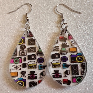 May include: A pair of silver teardrop-shaped earrings with a white background and colorful camera and video camera designs.