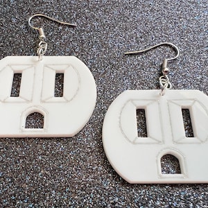 May include: A pair of white, plastic earrings shaped like electrical outlets. The earrings have silver hooks.