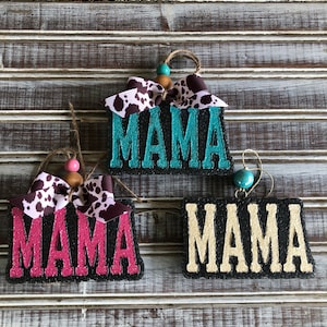 May include: Three black wooden signs with the word "MAMA" written in different colors. The signs are decorated with brown and white cow print bows and wooden beads.