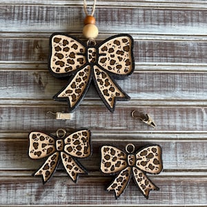 May include: Three wooden bow ornaments with a leopard print design. The largest bow is hanging from a string with wooden beads. The two smaller bows are on a wooden surface.