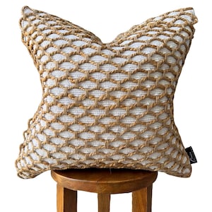 May include: Square decorative pillow with a woven pattern in natural and off-white tones. The pillow has a textured, geometric design, and is placed on a small wooden stool. The pillow's tag is visible on the lower right corner.