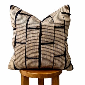 May include: Square decorative pillow with a beige woven fabric and a bold black geometric pattern. The pillow is placed on a small wooden stool, showcasing its design and texture. The pillow's design features a grid-like pattern.