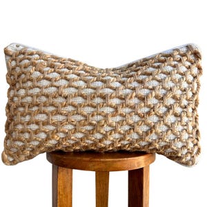 May include: A rectangular decorative pillow with a woven pattern. The pillow features a natural color scheme with a beige and off-white design. The pillow is placed on a wooden stool.