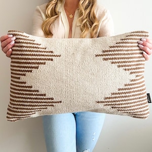 May include: A rectangular cushion in beige with brown geometric patterns. The cushion has a woven texture and a neutral colour palette, suitable for various home decor styles. The brown pattern is a series of horizontal lines and zigzags.
