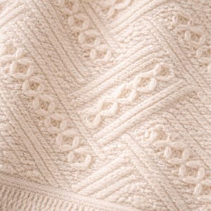May include: Close-up of a cream-colored knitted blanket with a detailed pattern. The design features a combination of textured stitches, including cable and diamond patterns, creating a cozy and intricate texture.