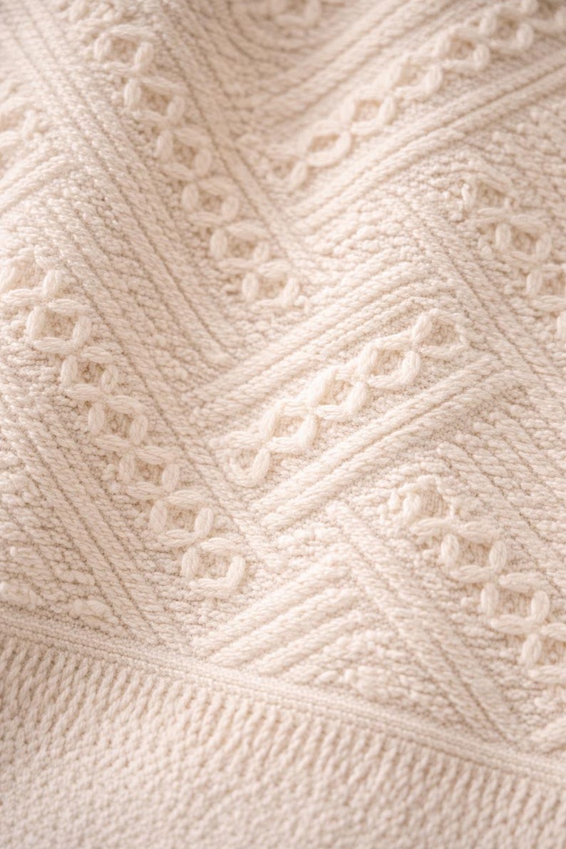 May include: Close-up of a cream-colored knitted blanket with a detailed pattern. The design features a combination of textured stitches, including cable and diamond patterns, creating a cozy and intricate texture.
