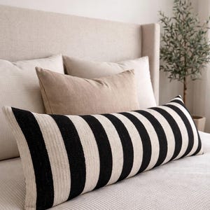 Pode incluir: Uma almofada longa e retangular com listras pretas e brancas arrojadas. A almofada está colocada numa cama com outras almofadas de tons neutros e uma cabeceira bege. A estética geral é moderna e minimalista.