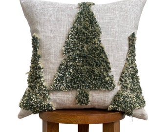 May include: A square decorative pillow with a neutral linen-like background. Three Christmas tree designs are embroidered on the front with a textured, shaggy green and tan yarn. The pillow is placed on a wooden stool.