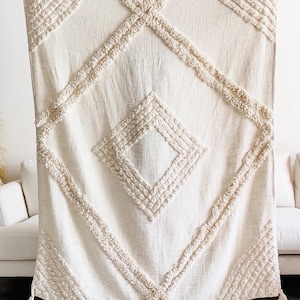 Puede incluir: Manta tejida de color crema con un patrón de diamantes. La manta presenta formas de diamantes texturizadas en relieve y detalles de flecos. El diseño es simétrico, con un diamante grande en el centro y diamantes más pequeños en las esquinas. Borlas cuelgan de la parte inferior.