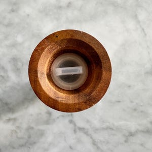 May include: Top view of a wooden salt or pepper grinder with a clear plastic lid. The lid has a small opening for dispensing salt or pepper.