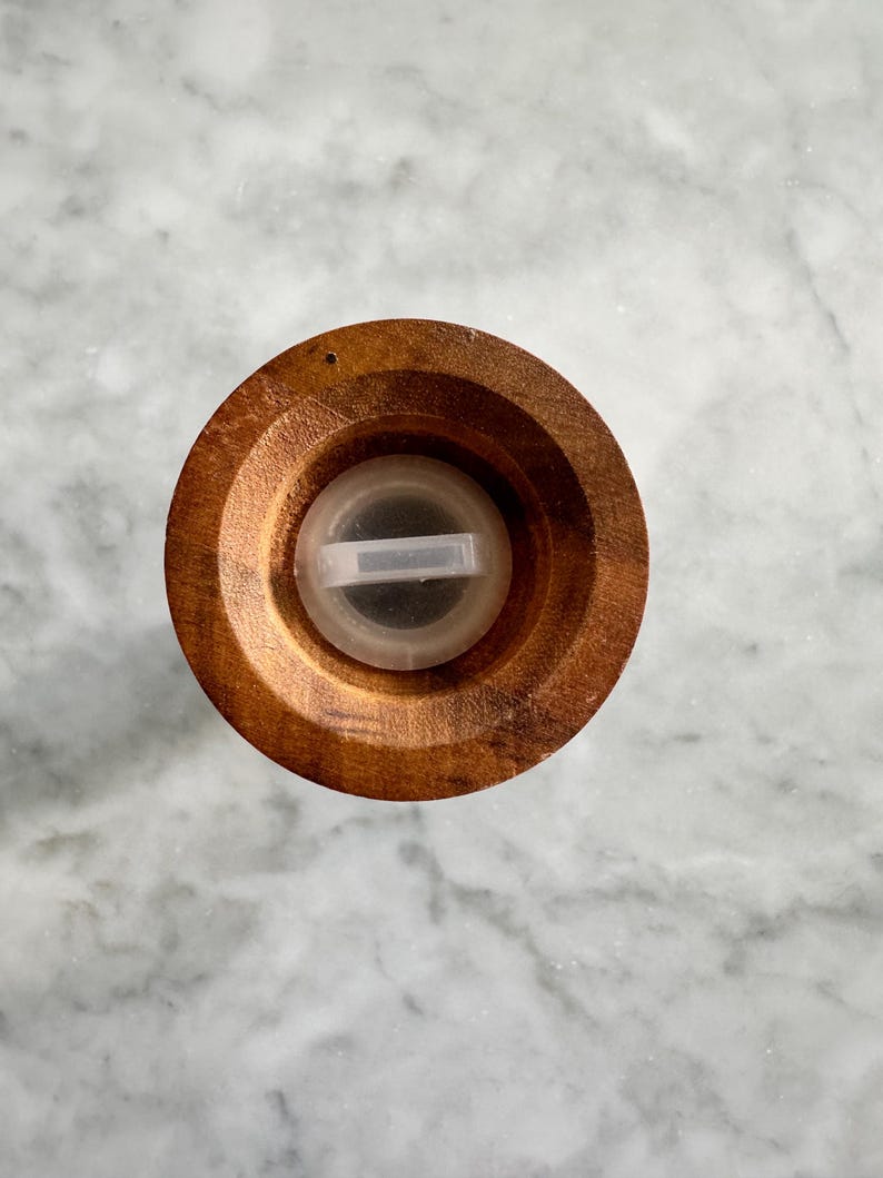 May include: Top view of a wooden salt or pepper grinder with a clear plastic lid. The lid has a small opening for dispensing salt or pepper.