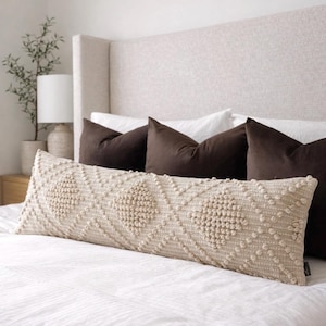 Könnte beinhalten: Ein langes, rechteckiges Lendenkissen in hellem Beige mit einem strukturierten Rautenmuster aus erhabenen, getufteten Knoten. Das Kissen liegt horizontal auf einem Bett vor drei einfarbigen, dunkelbraunen, quadratischen Kissen.