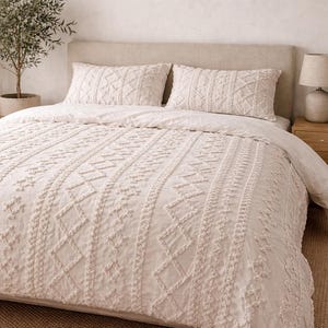 Pode incluir: Conjunto de roupa de cama creme com um padrão texturizado em relevo. O conjunto inclui uma capa de edredão e duas fronhas. O design incorpora elementos geométricos e lineares, criando uma estética acolhedora. A cama está sobre um fundo neutro.